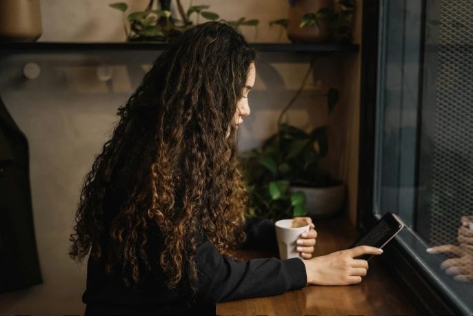 A woman with long curly hair sits by a window, holding a coffee cup while reading on an e-reader device in a cozy café setting.